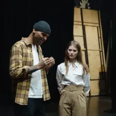 Mężczyzna w czapce i koszuli w kratę rozmawia z kobietą na scenie. Stoją blisko siebie, przygotowując się do występu.