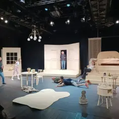 Scena teatralna z ludźmi leżącymi na podłodze i siedzącymi na schodach. Czy to **farsa, co to** za zamieszanie?