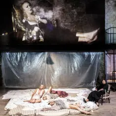 Na scenie teatralnej, w tle projekcja kobiety z fajką, a na dole leżące kobiety. Aluzja do przeszłości.