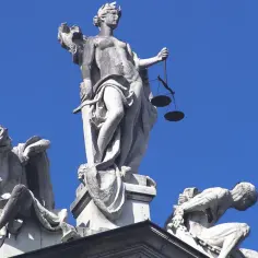 Kamienna Temida z wagą i mieczem, otoczona innymi postaciami, symbolizuje sprawiedliwość na tle błękitnego nieba.