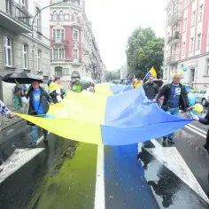 Ludzie niosą wielką flagę w barwach Ukrainy. W deszczu idzie grupa, wśród nich młoda kobieta, która nie jest gorolem.