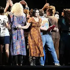 Scena z musicalu. Grupa ludzi na scenie, w centrum kobieta w sukience w kwiaty.