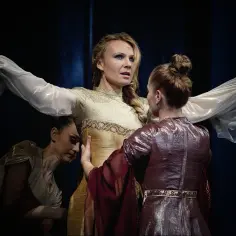 Anna Guzik w złotej sukni, otoczona przez dwie tancerki w teatralnych strojach, tworzy scenę pełną emocji.
