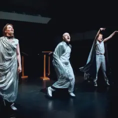Aktorzy na scenie, w srebrnych strojach, odgrywają scenę pełną emocji, być może symbolizującą śmierć.