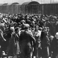 Tłum ludzi w pasiastych ubraniach i cywilnych strojach stoi przed rzędem wagonów towarowych.