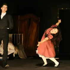 Mężczyzna w czarnym fraku i dziewczynka w czerwonej sukience na scenie. W tle drewniana szafa i łóżko. Całość tworzy teatralny kontekst.