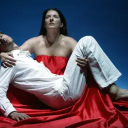 Kobieta w czerwonej draperii trzyma na rękach mężczyznę w białej koszuli. Dramatyczna scena z performance'u.
