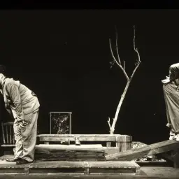 Dwóch mężczyzn w kapeluszach na scenie, w scenografii przypominającej teatr absurdu. Jeden szuka czegoś, drugi stoi na chybotliwej belce.