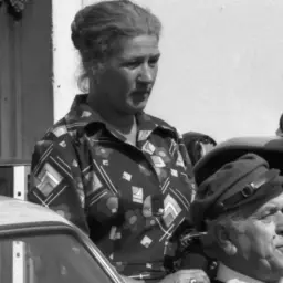 Maria Zbyszewska, wpatrzona w dal, obok mężczyzny w czapce. W tle traktor i samochód.