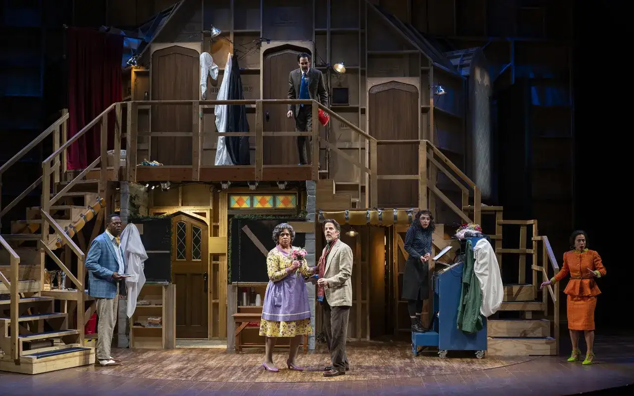Scena teatralna z ludźmi w dziwacznych strojach, drewnianymi schodami i drzwiami. Czy to farsa? Co to za chaos!