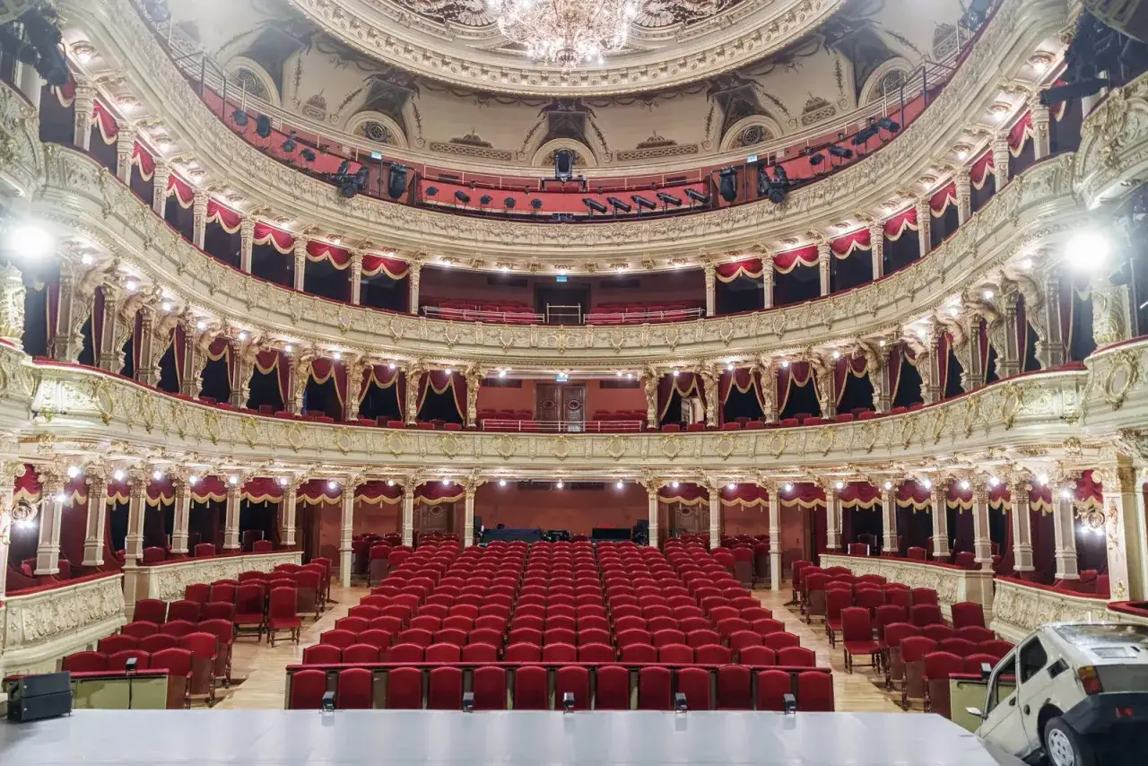Wnętrze teatru z rzędami czerwonych foteli i bogatymi zdobieniami. Kontrapunkt klasyki i nowoczesności, gdzieś na widowni stoi Fiat 126p.