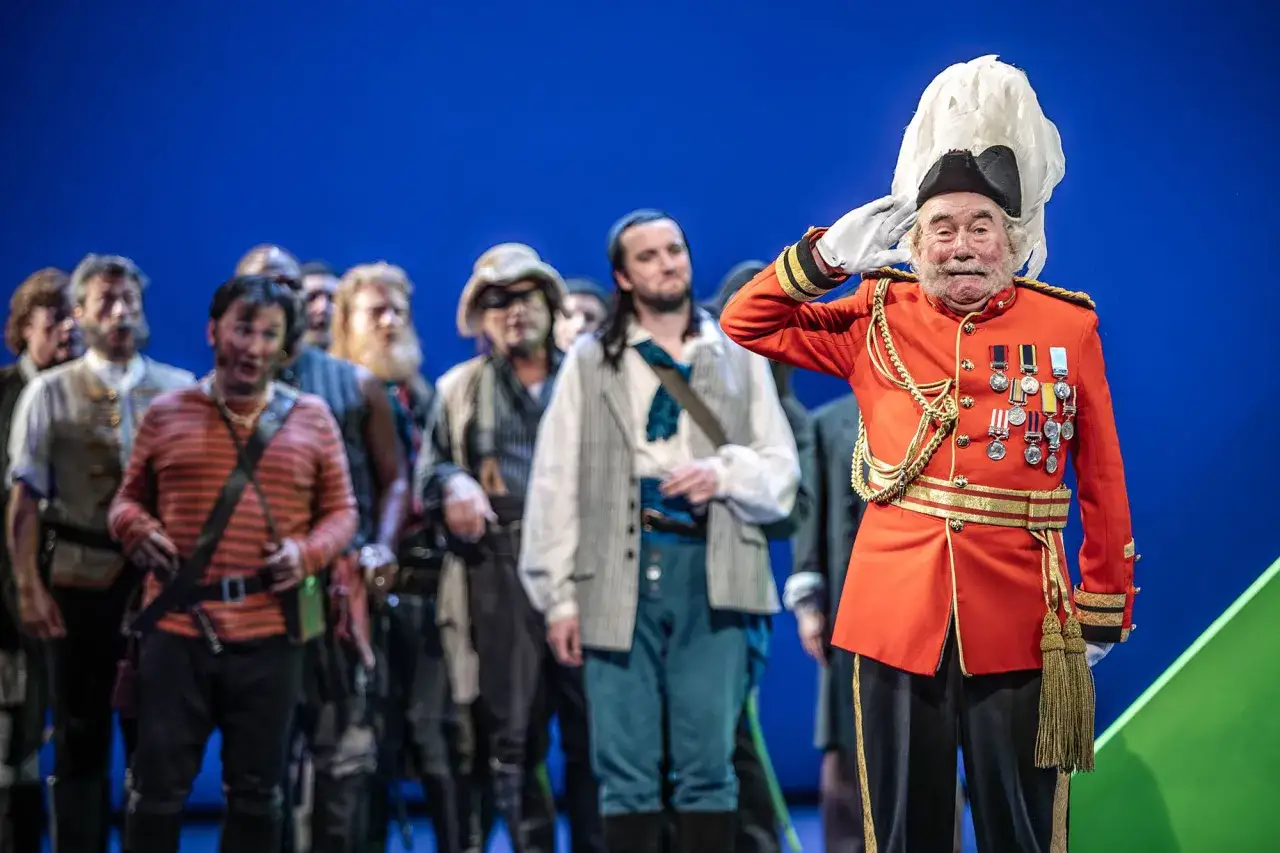 Starszy mężczyzna w mundurze, ozdobionym medalami, salutuje na scenie. W tle grupa aktorów w kostiumach, przygotowana do występu w operetce.