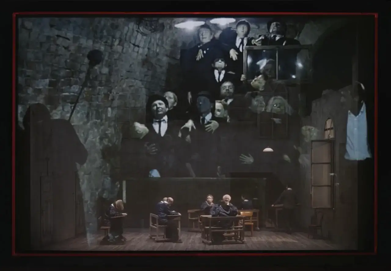 Scena teatralna z projekcją postaci w melonikach i białych koszulach. Na dole siedzą uczniowie przy ławkach. To sugestywna aluzja do przeszłości.