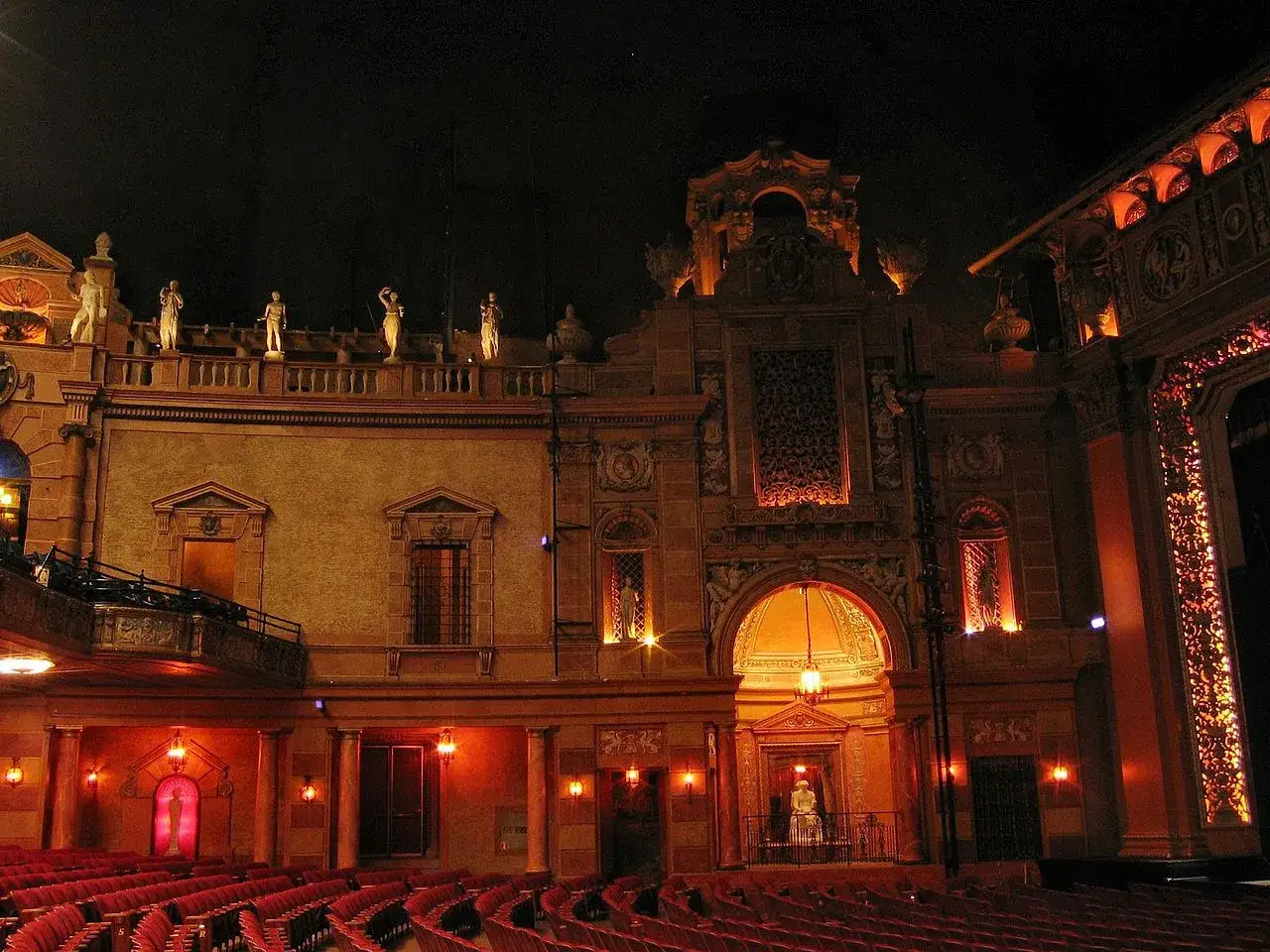 Wnętrze teatru z czerwonymi fotelami i bogatą scenografią. Widok na zdobioną fasadę, rzeźby i łukowate wejście.