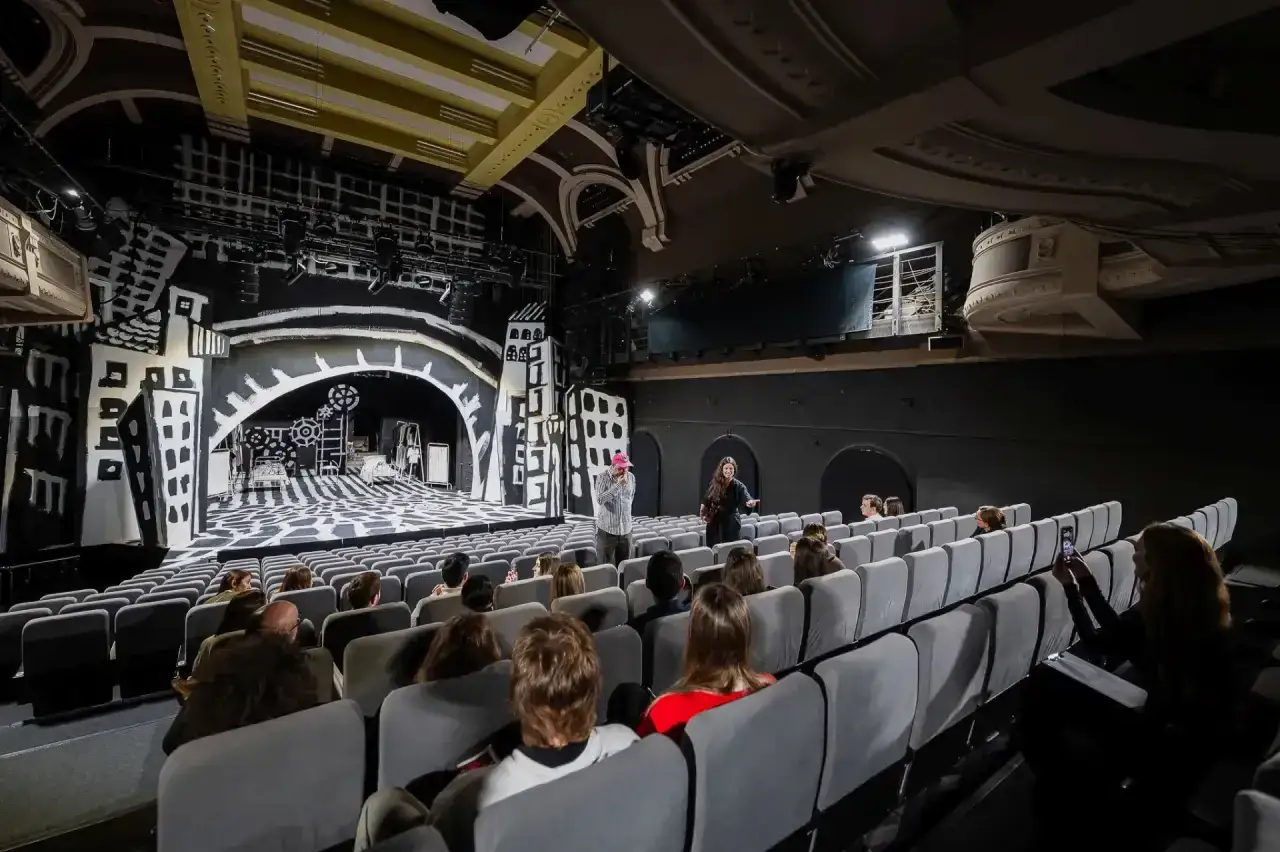Widownia teatru z widokiem na scenę w stylu ekspresjonistycznym. Grupa widzów, wśród nich osoba robiąca zdjęcie telefonem, czeka na rozpoczęcie spektaklu, który zapowiada się jak ciekawy felieton wizualny.