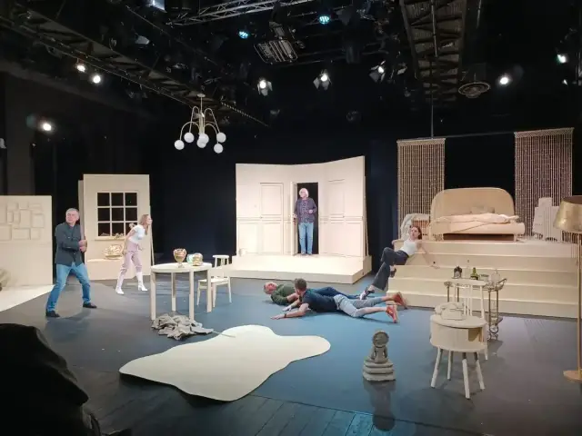 Scena teatralna z ludźmi leżącymi na podłodze i siedzącymi na schodach. Czy to **farsa, co to** za zamieszanie?